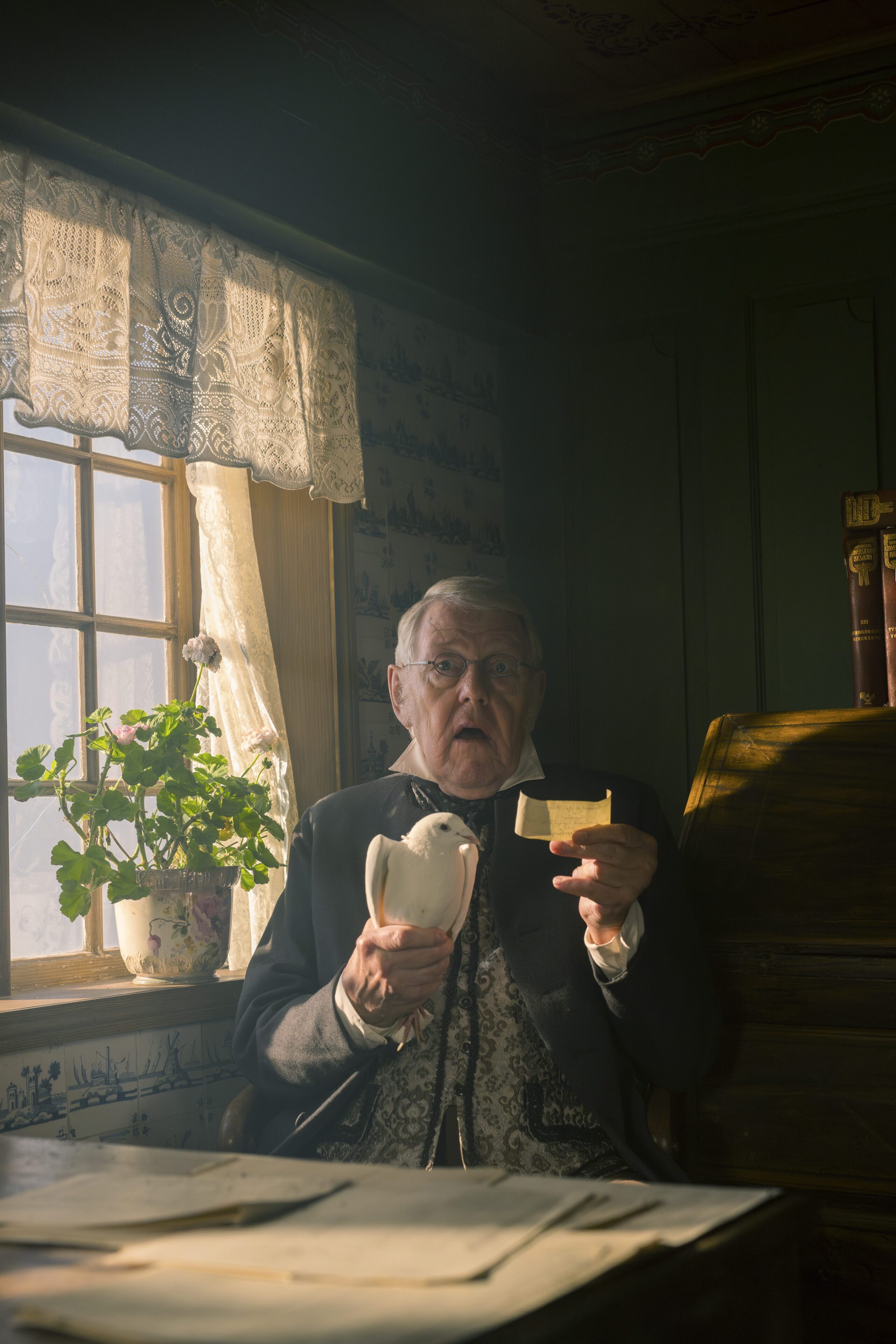 A vertical portrait of an elderly man with glasses in a historic, dimly lit room. He looks startled with his mouth open while holding a white pigeon in one hand and a piece of paper in the other next to a sunny window.