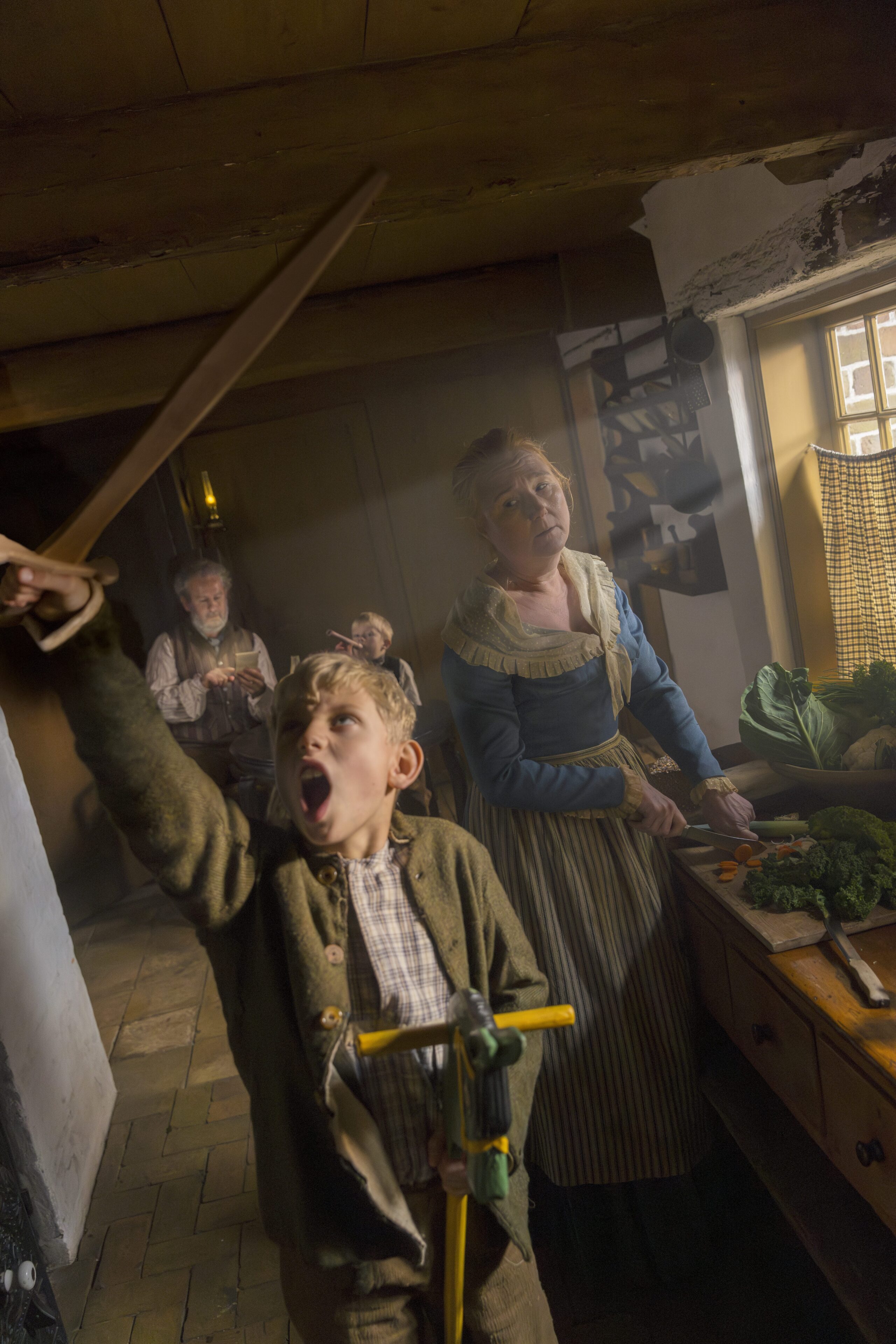 A dynamic, low-angle shot in a historic kitchen setting. In the foreground, an enthusiastic young boy shouts while raising a wooden toy sword. Behind him, a woman in historical attire continues her meal preparation at a wooden counter.