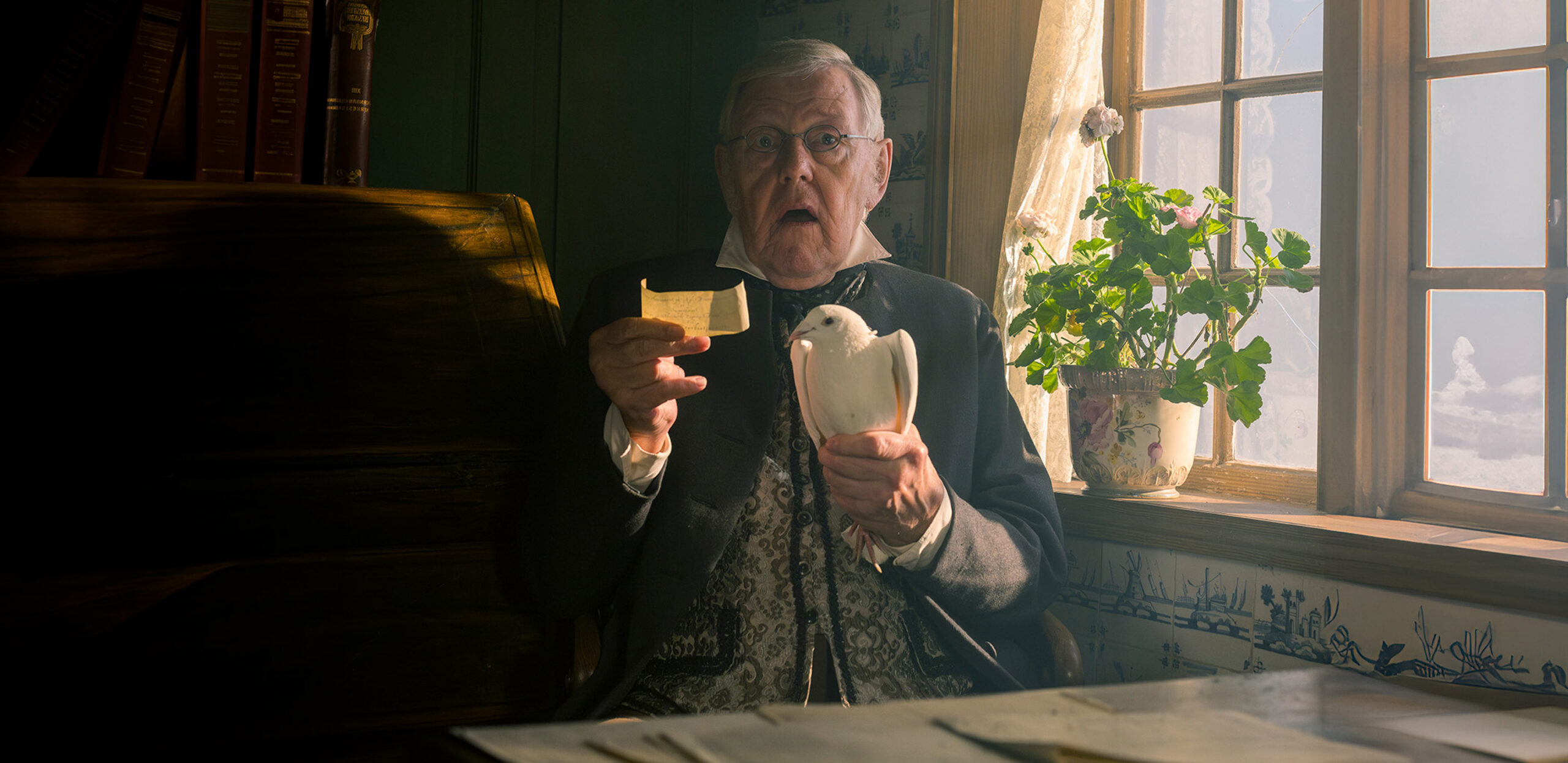 A wide horizontal view of an elderly man with glasses in historical attire, looking shocked with his mouth agape. He is holding a white pigeon and a small slip of paper while sitting next to a bright window with a potted plant.