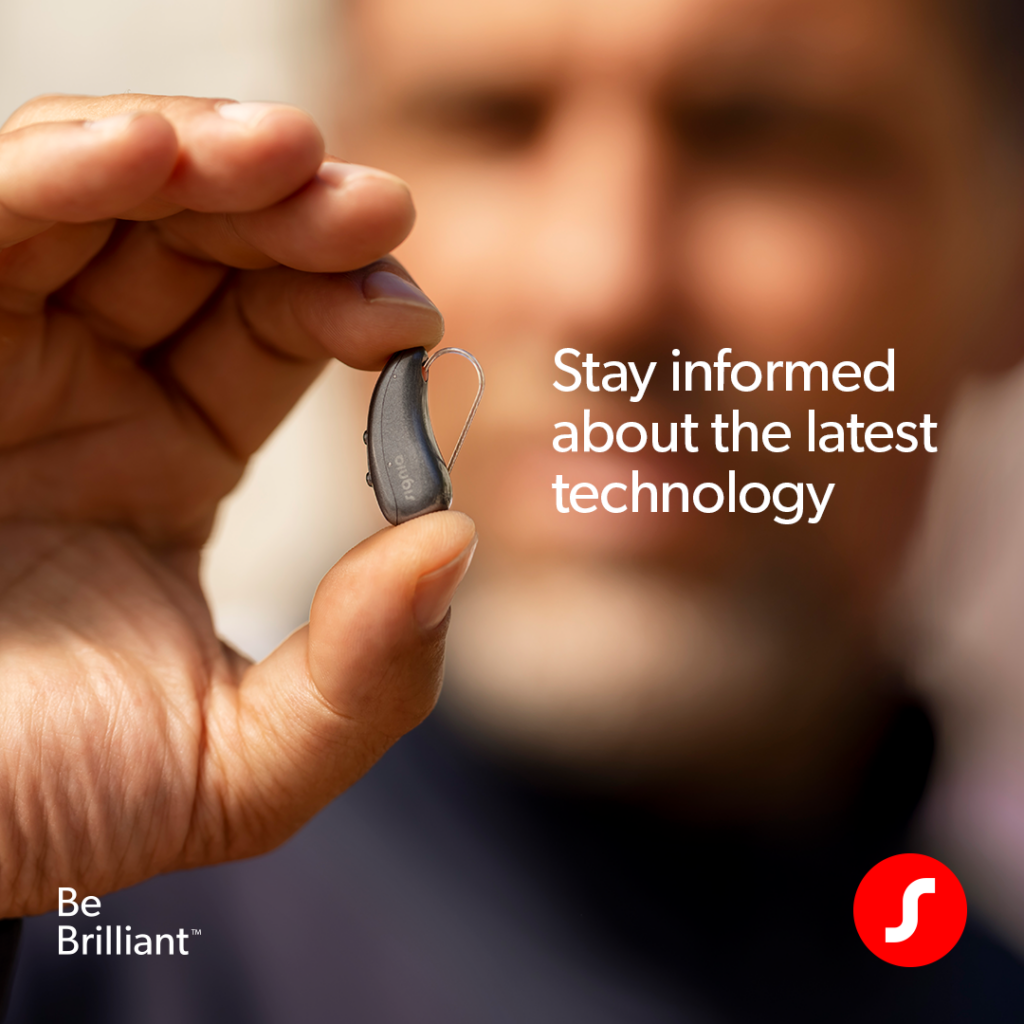 A close-up of a hand holding a tiny, sleek grey Signia behind-the-ear hearing aid between the thumb and forefinger. A man's face is softly blurred in the background. Text reads, 'Stay informed about the latest technology' next to the product. A Signia logo in the lower right corner.