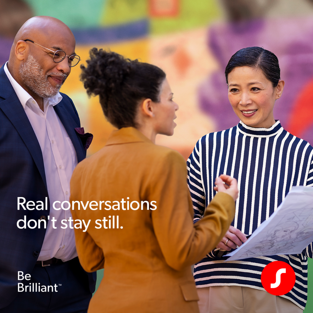 A medium shot of three people in conversation against a blurred, colorful mural background. A woman in a striped turtleneck holds a blueprint while talking to a woman in an amber blazer and a man in a navy suit. Text reads, 'Real conversations don't stay still.' A Signia logo in the lower right corner.