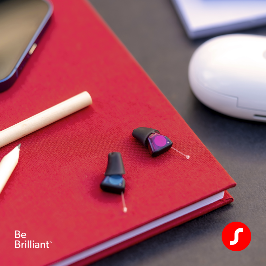 A close-up shot of two tiny in-the-ear hearing aids—one with a blue faceplate and one with a magenta faceplate—resting on a textured red notebook alongside a white pencil. A white charging case is blurred in the background, with 'Be Brilliant™' and Signia logo in the bottom corner.