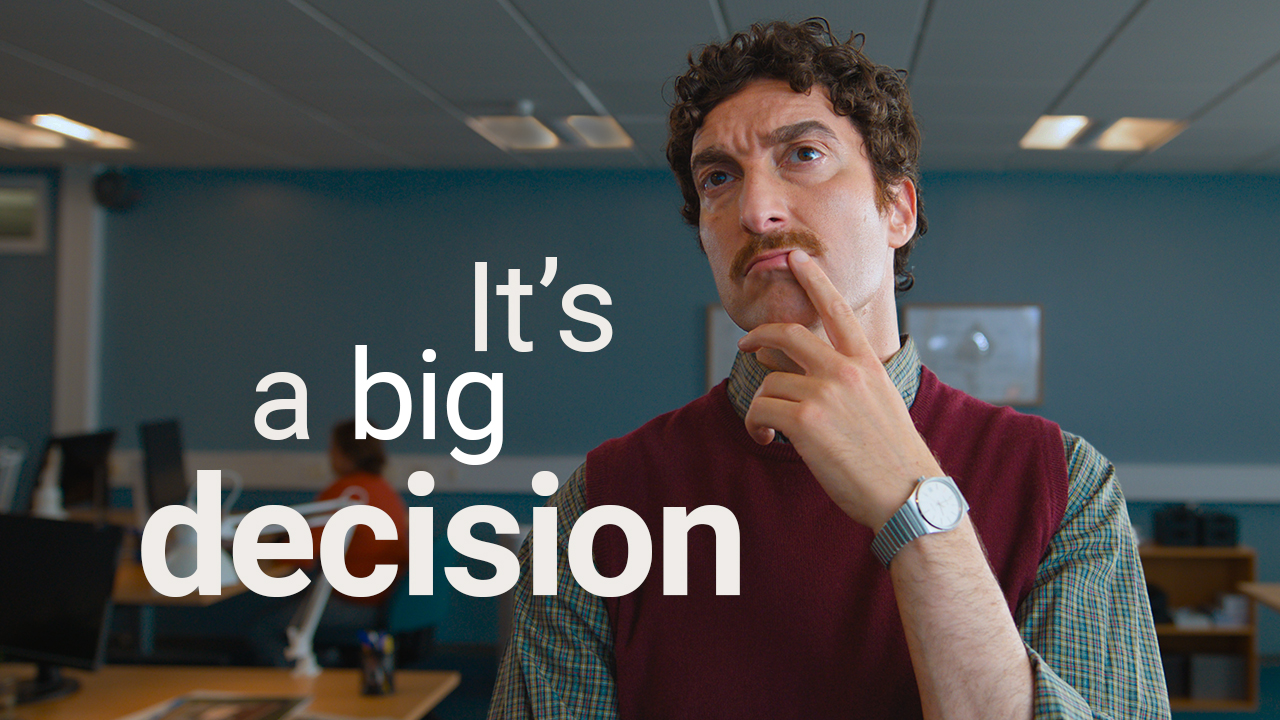 A medium shot of a man with a mustache and a maroon vest looking upwards thoughtfully with his finger on his chin. Large white text overlays the office background, reading: 'It's a big decision'.