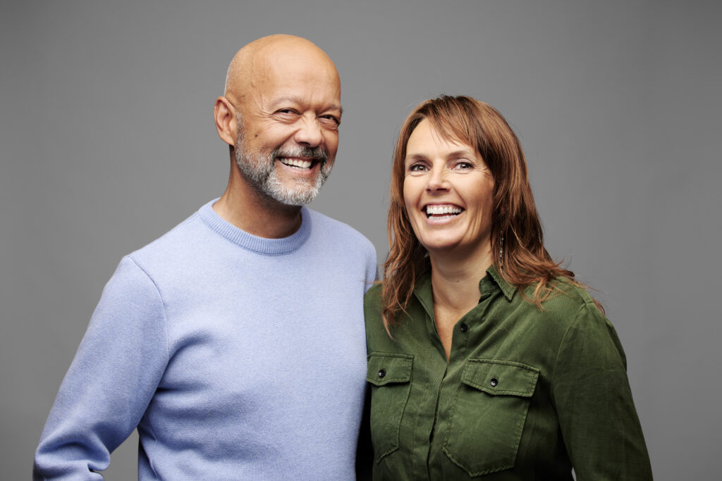 A portrait of a smiling couple standing together against a grey backdrop. The man on the left is bald with a grey beard and is wearing a light blue sweater. The woman on the right has brown hair and is wearing an olive green button-up shirt.