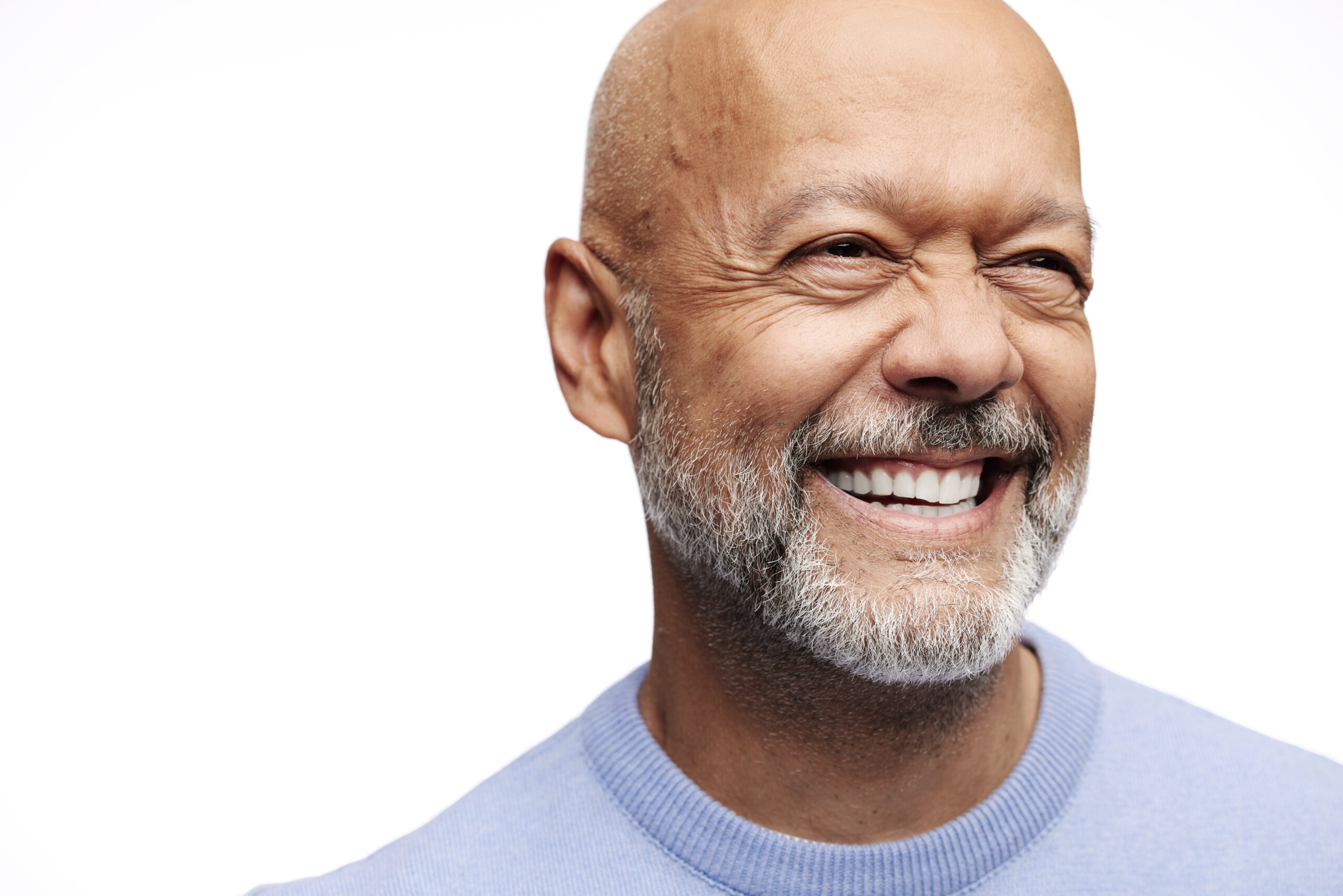 A tight, high-contrast portrait of a bald man with a grey beard, smiling broadly with clean white teeth. He is wearing a light blue sweater and looking off-camera to the right against a stark white background.