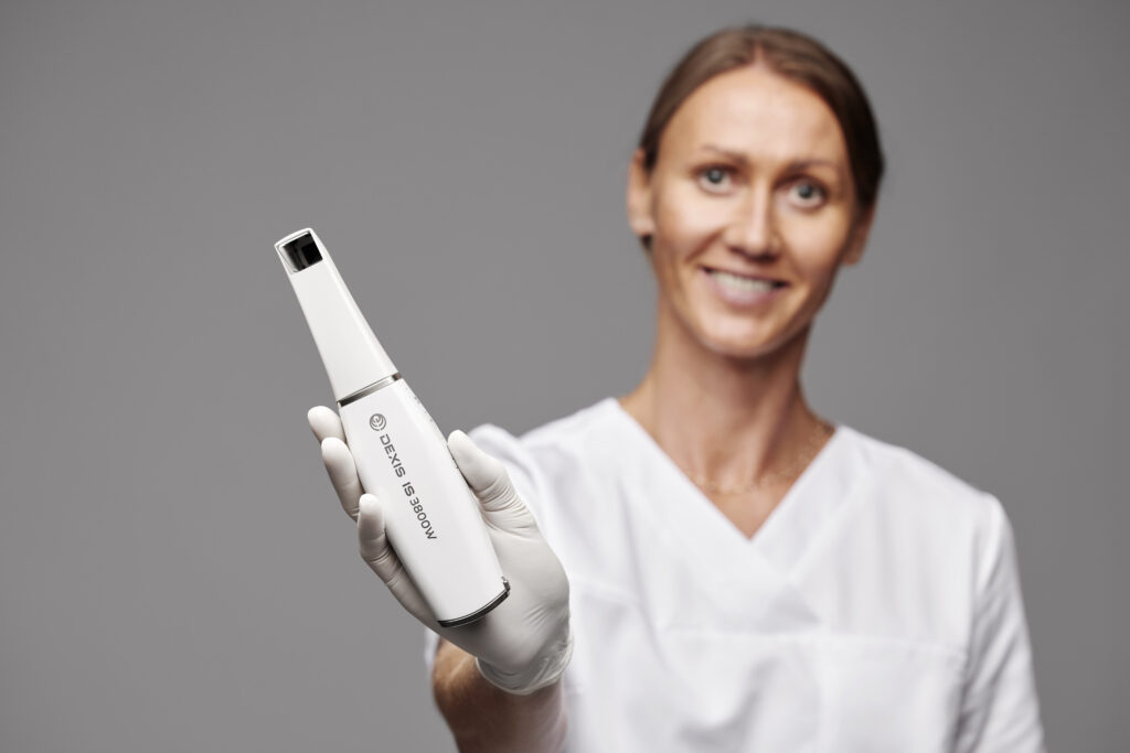 A close-up shot focused on a gloved hand holding a white 'DEXIS IS 3800W' intraoral dental scanner toward the camera. A dental professional in white scrubs is visible but softly blurred in the background against a grey wall.