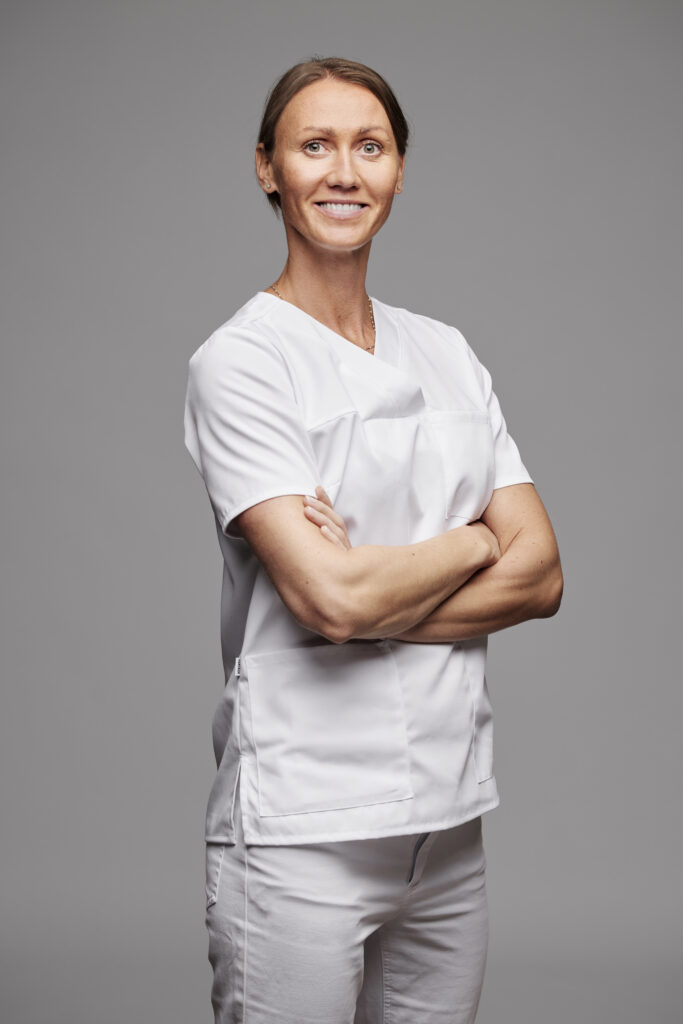 A studio portrait of a tall woman with straight brown hair tied back, wearing white medical scrubs and standing with her arms crossed. She is looking directly forward with a confident, pleasant smile against a neutral grey background.