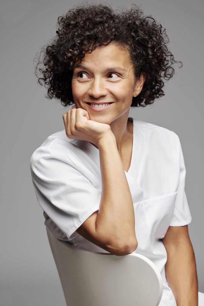 A studio portrait of a woman with short, curly black hair and a white dental tunic, resting her chin on her hand while leaning over the back of a white chair. She is looking away to the side with a gentle, curious smile against a grey background.