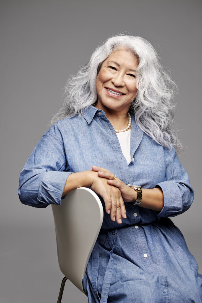 A portrait of the same older woman with wavy white hair, now sitting in a white chair and looking directly at the camera with a warm, pleasant smile. She is wearing a blue denim shirt and a pearl necklace against a solid grey background.
