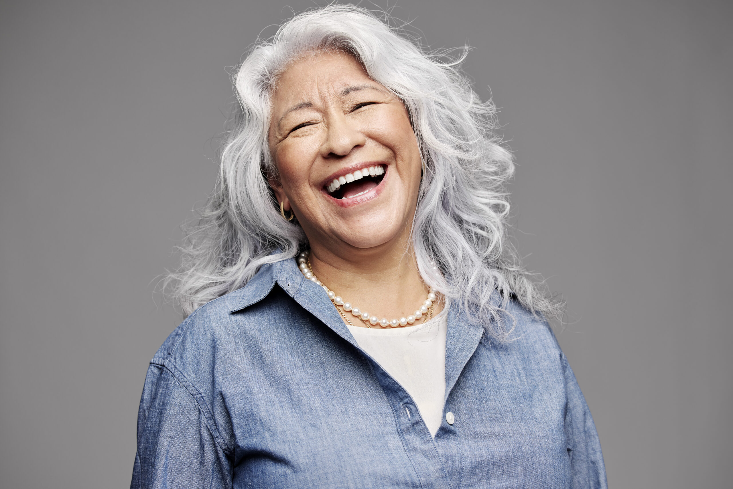 A close-up studio portrait of an older woman with long, voluminous grey-and-white wavy hair. She is wearing a blue denim button-up shirt and a white pearl necklace, laughing with a joyful, wide-open smile against a solid grey background.