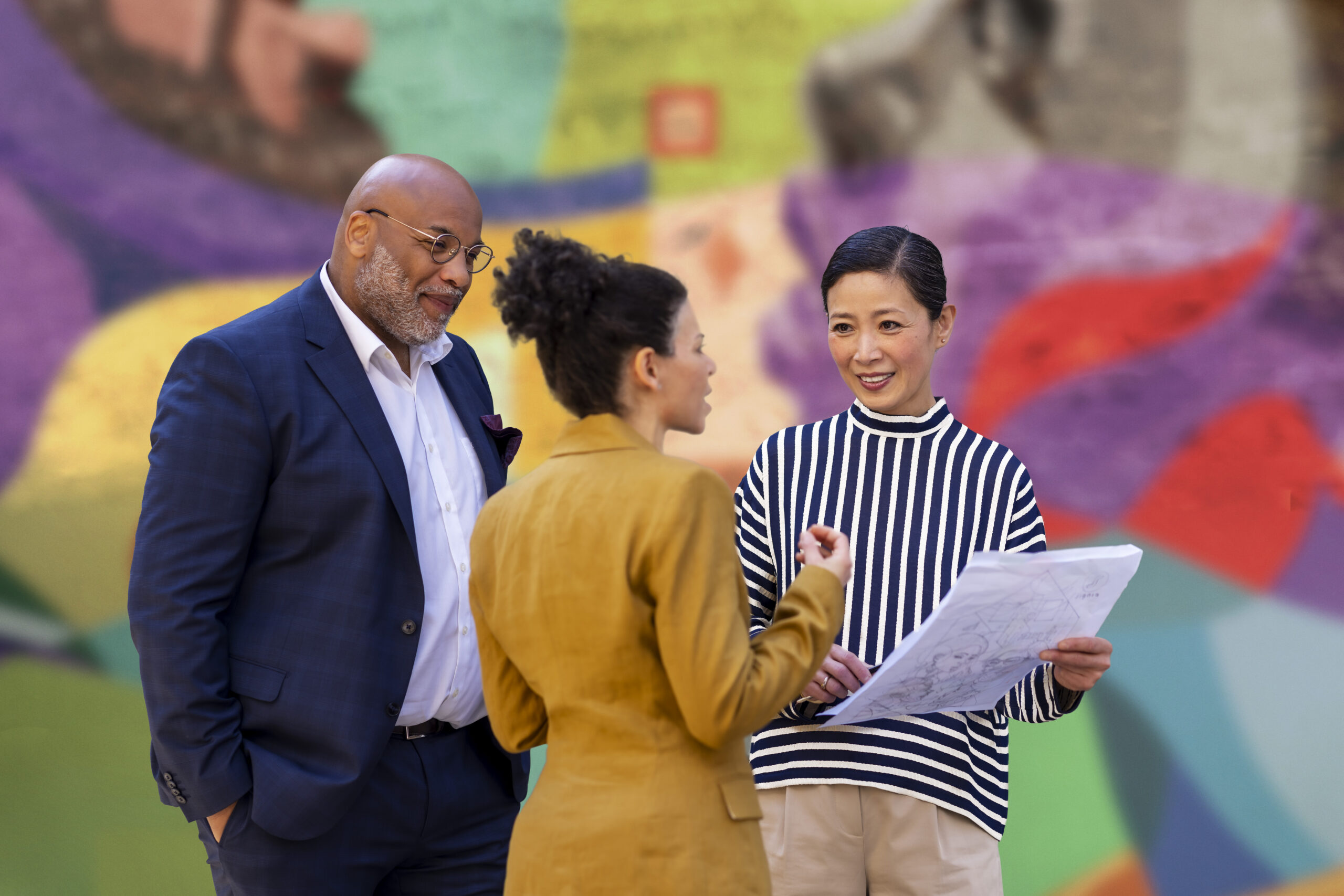 A medium shot of a group of three professionals - a man in a navy suit, a woman with her hair in a bun wearing an amber blazer, and a woman in a black-and-white striped turtleneck—reviewing an architectural drawing outdoors in front of a vivid, multicolored abstract mural.