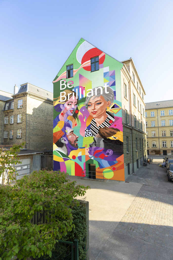A wide shot of a five-story urban building featuring a massive, vibrant, multi-colored painted mural of diverse faces talking. The text 'Be Brilliant' is written in large white letters across the top section of the artwork.