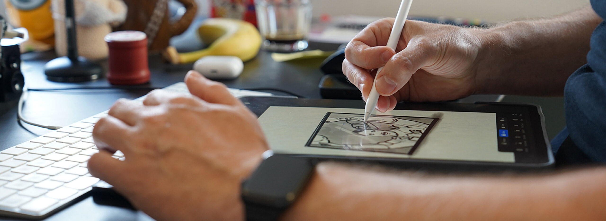 Close-up of a person's hands using a digital pen to draw an illustration on a tablet next to a computer keyboard.