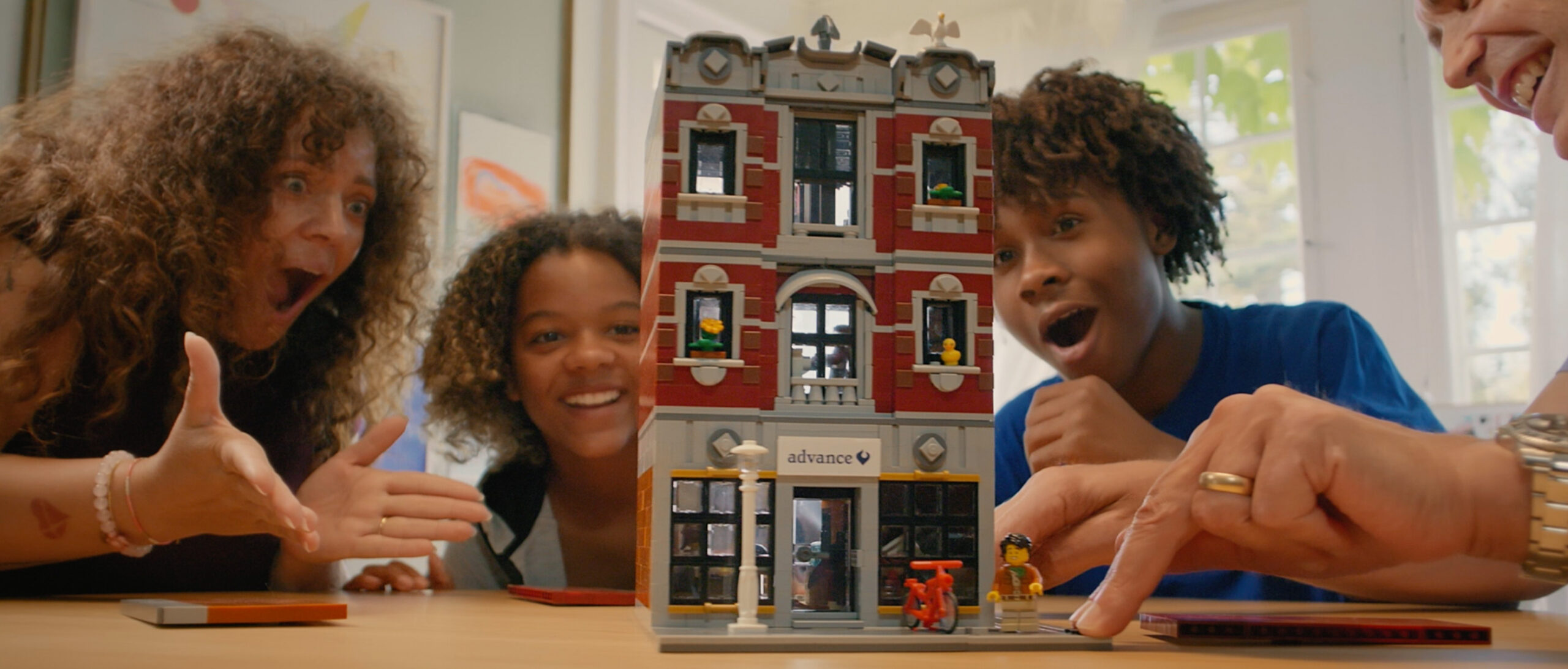 Two excited children and two adults looking with wide eyes and open mouths at an intricate multi-story LEGO building model on a table. The building has an Advance logo above the entrance.