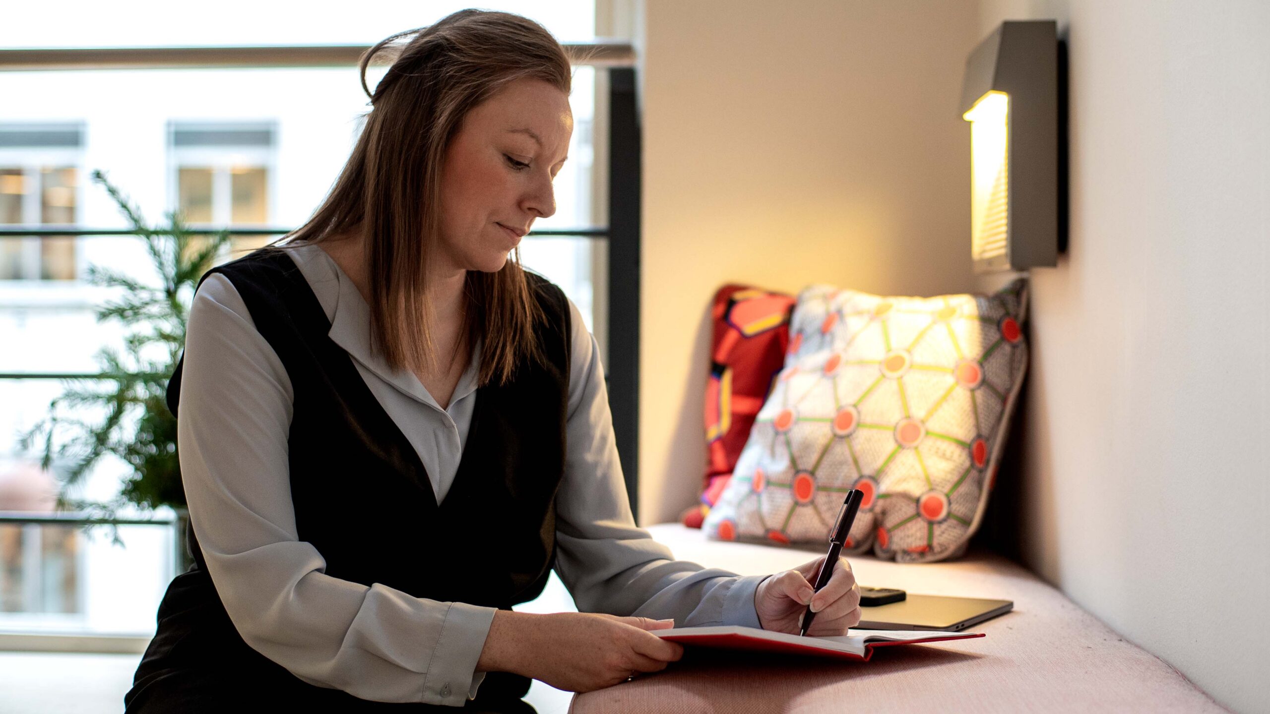 woman sitting on a cushioned window bench, wearing a light grey blouse and a black vest, focused on writing in a notebook with a pen.