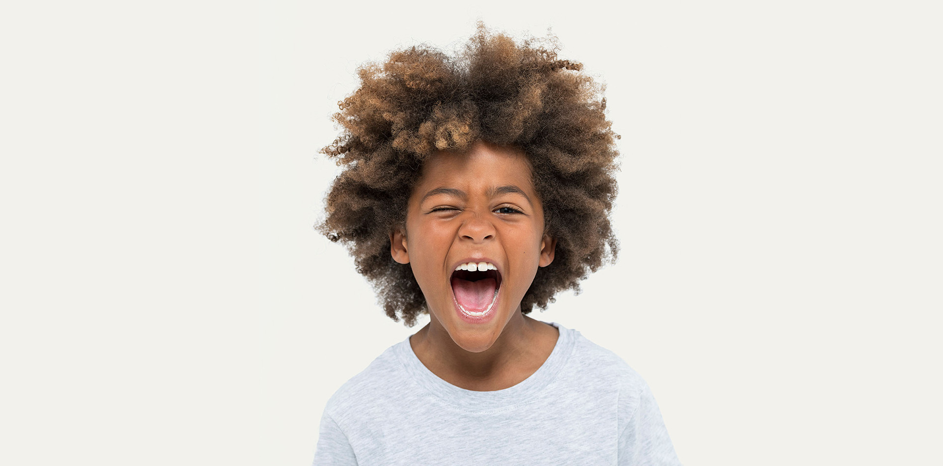 A wide portrait of a young boy with an afro hairstyle wearing a plain light grey T-shirt, making a funny face by winking one eye and opening his mouth wide to shout playfully against a clean light grey background.