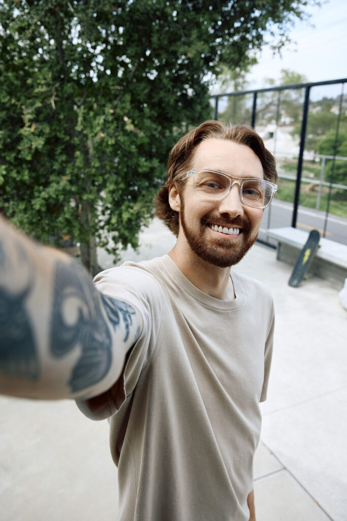 A high-angle selfie of a smiling man with a beard, clear-framed glasses, and a tattooed arm extending toward the camera. He is wearing a beige t-shirt, and a skateboard is visible on the ground behind him.
