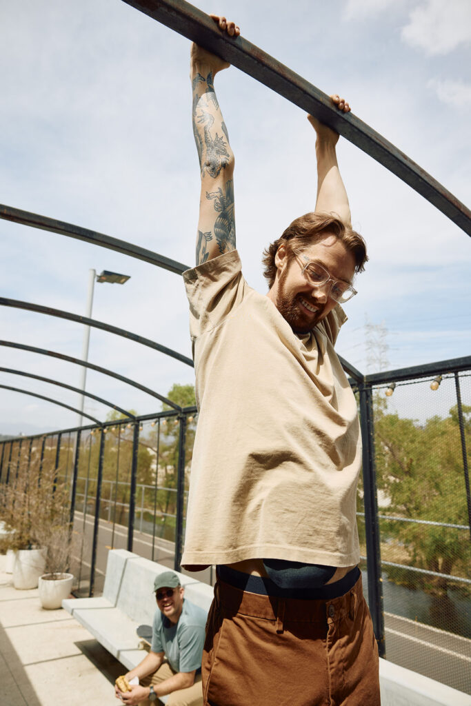 An outdoor shot of a man hanging playfully from an overhead metal bar structure, causing his beige t-shirt to lift and reveal the top of a black ostomy pouch at his waistline. Another man is seated on a bench in the background.