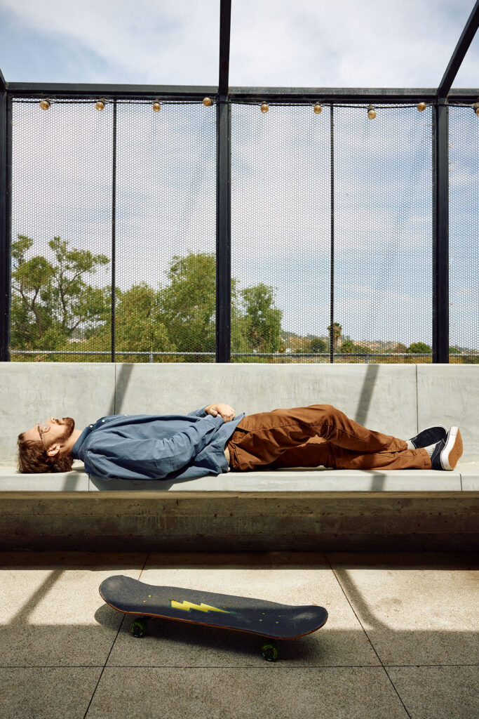 A man in a blue button-up shirt and brown pants lies flat on his back resting on an outdoor concrete bench. On the ground below the bench sits a black skateboard with a yellow lightning bolt design.