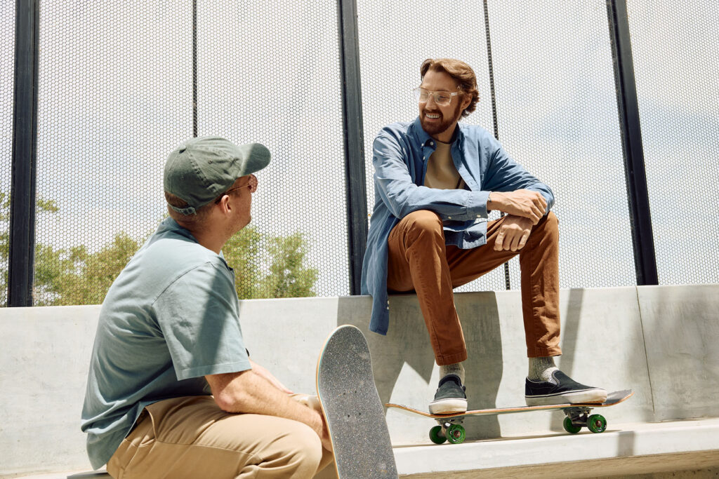 Two men chatting at an outdoor skatepark. On the right, a man with clear glasses and a blue button-up shirt sits on a concrete ledge with his feet resting on a skateboard. The other man sits facing him in the left foreground wearing a green cap.