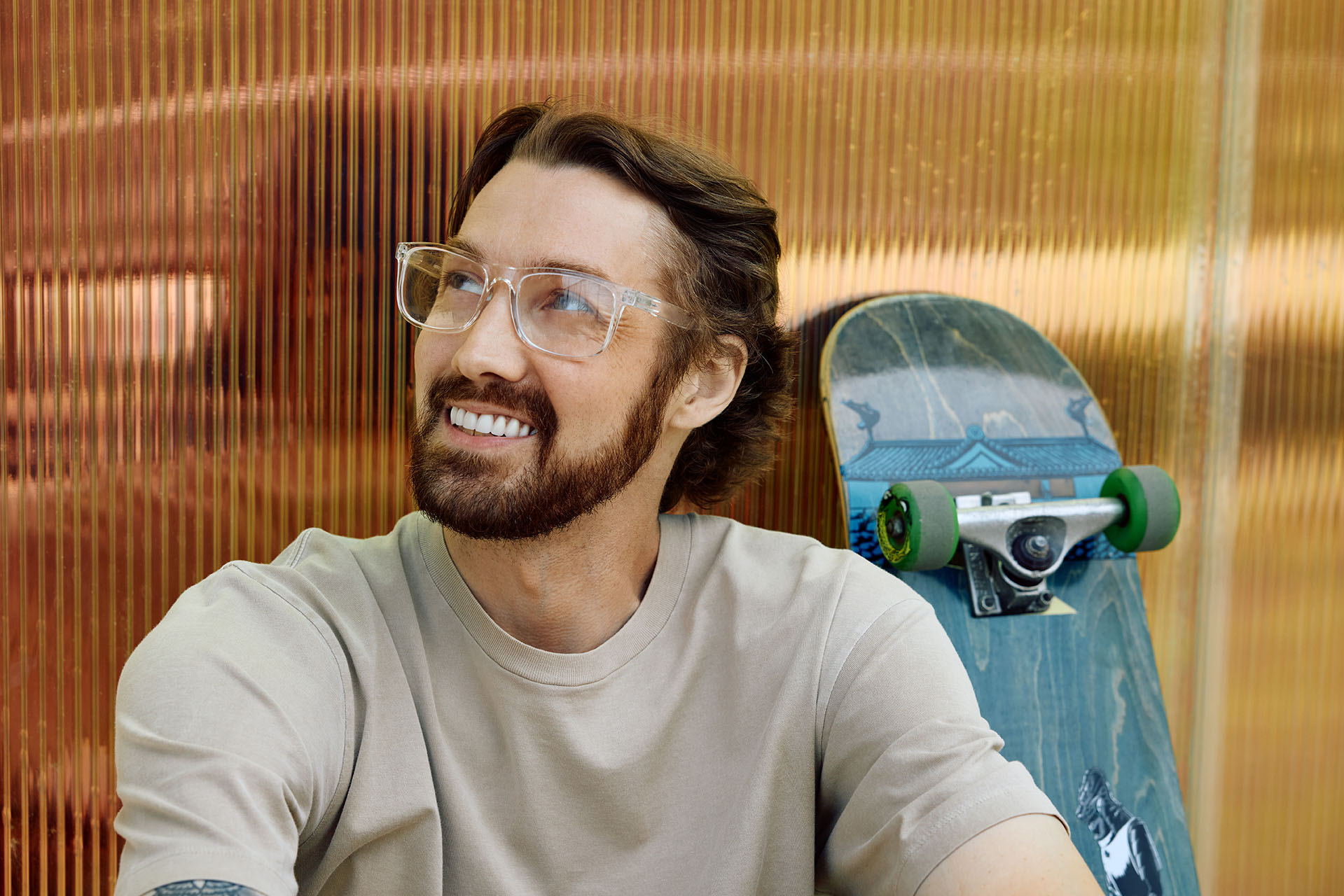 A close-up portrait of a smiling man with a beard and clear-framed glasses looking up and to the side. A blue skateboard is propped up behind him against a textured amber wall.