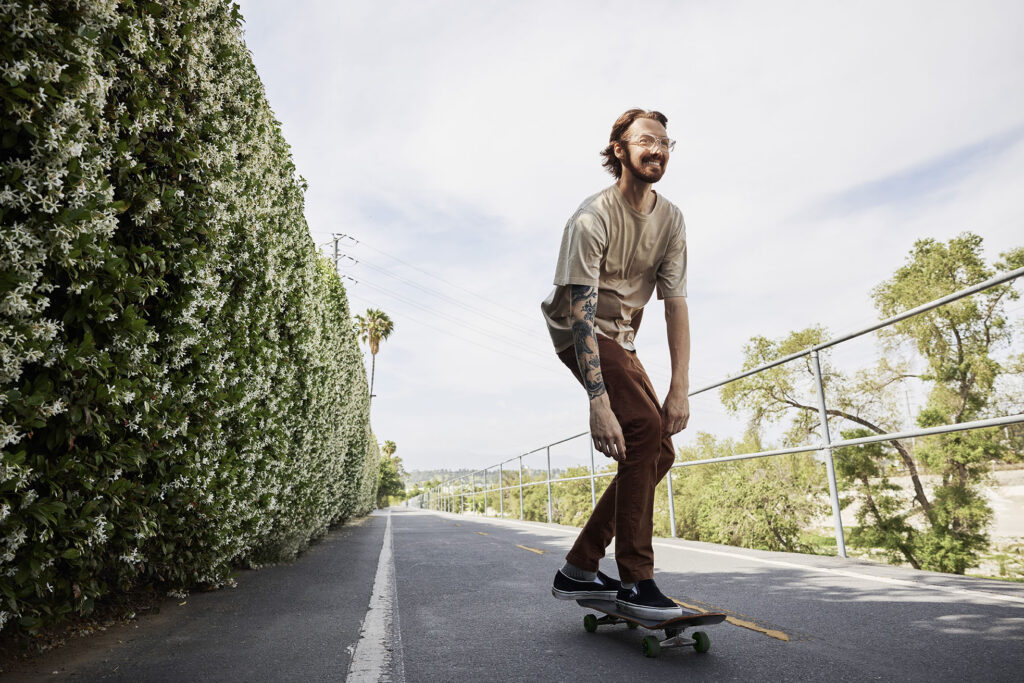 A man with a beard, tattoos, and glasses smiles as he rides a skateboard down an outdoor paved path lined with a tall, white-flowered green hedge on one side and a metal railing on the other.