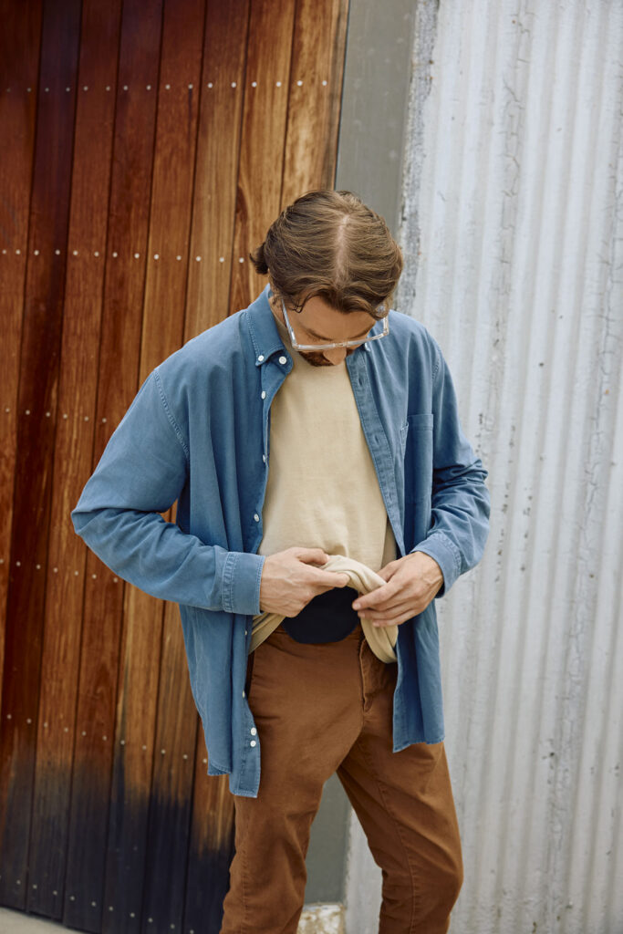A man with brown hair and clear-framed glasses stands outside against a wooden wall, lifting the hem of his beige t-shirt to look down at a black ostomy pouch tucked securely into his brown trousers.