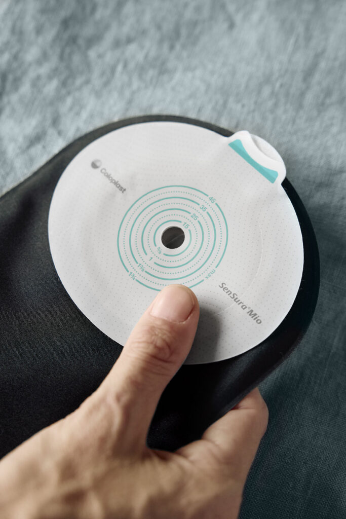A close-up shot of a hand holding a black ostomy pouch. The focus is on the white, circular adhesive barrier which features the Coloplast and SenSura Mio branding alongside green concentric measurement lines for custom cutting.