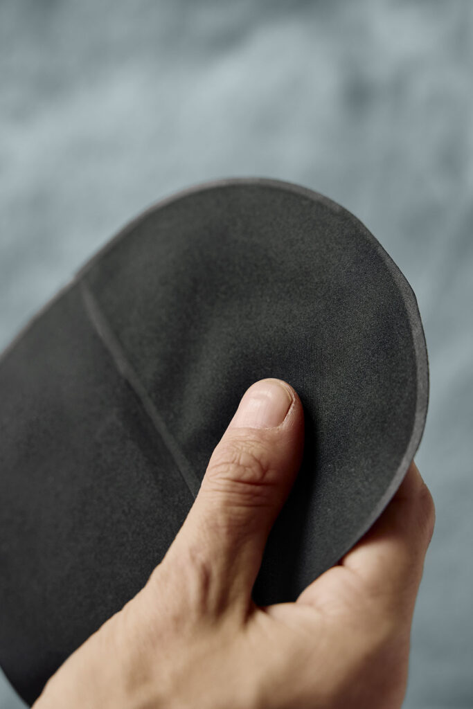 A close-up shot of a hand feeling the soft, textile material of a plain black circular ostomy pouch cover against a blurred grey background.