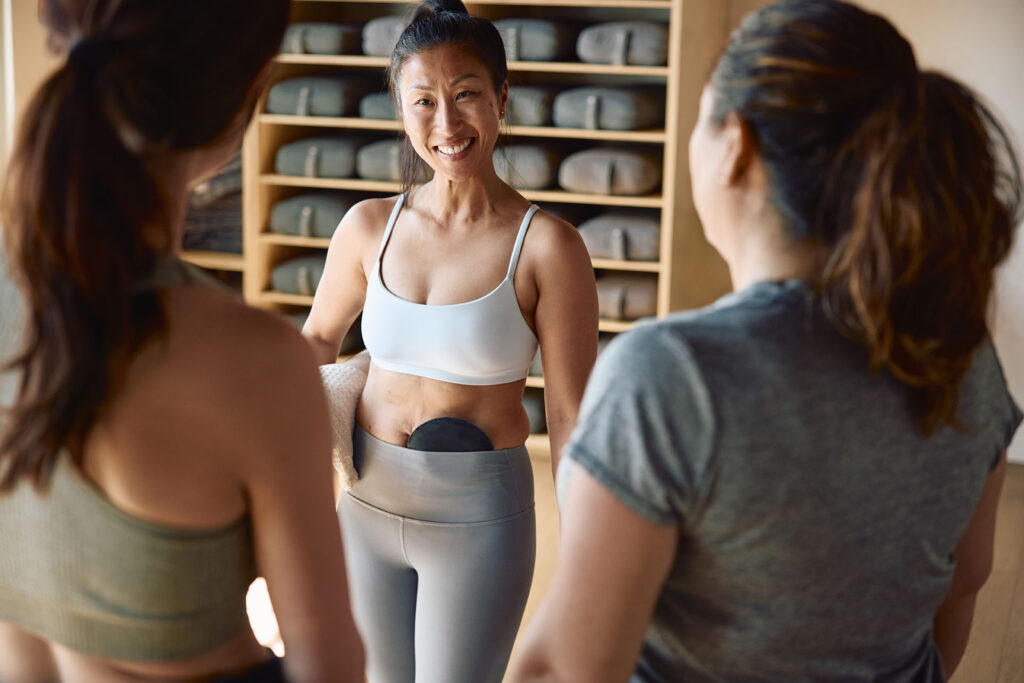 An East Asian woman in a white sports bra and grey leggings stands in a yoga studio, smiling warmly as she talks to two other women seen from behind. A black circular ostomy pouch cover is visible at her waist, and a shelf filled with yoga mats stands in the background.
