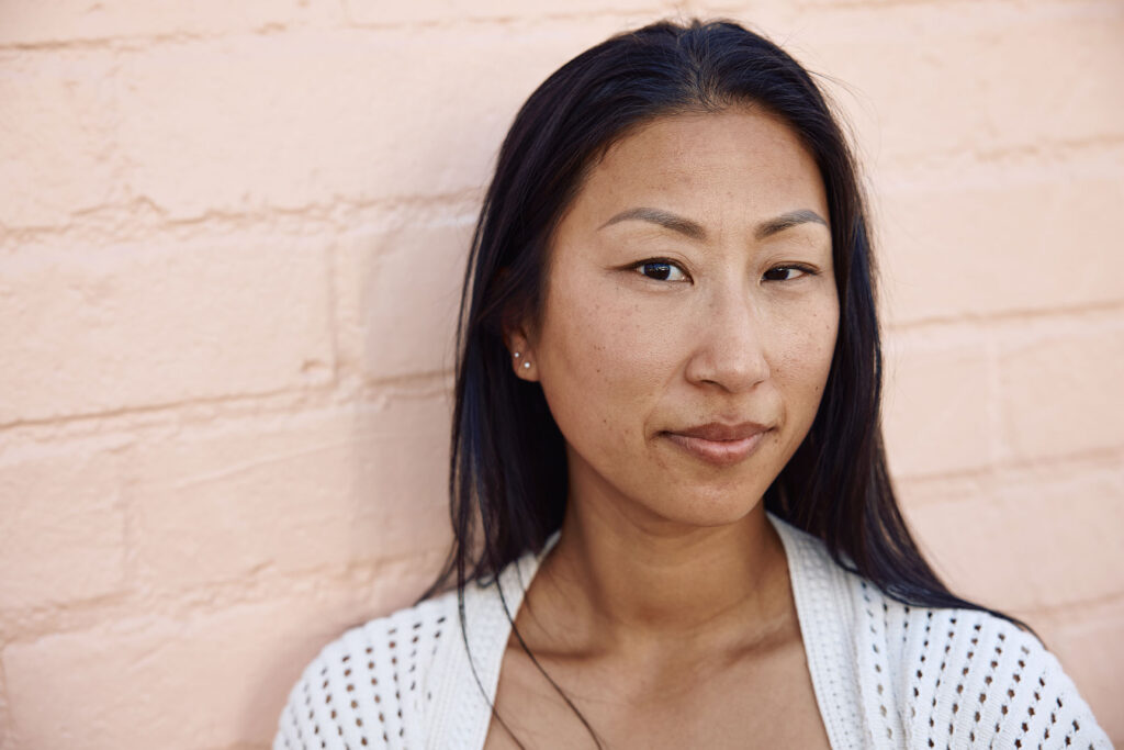 A close-up portrait of an East Asian woman with long dark hair, wearing a white crocheted top, looking directly at the camera with a gentle expression against a light pink brick wall.