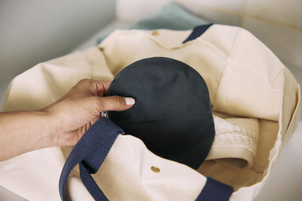 A top-down view of a person's hand pulling a black ostomy pouch out of a cream-colored canvas tote bag with dark blue straps.