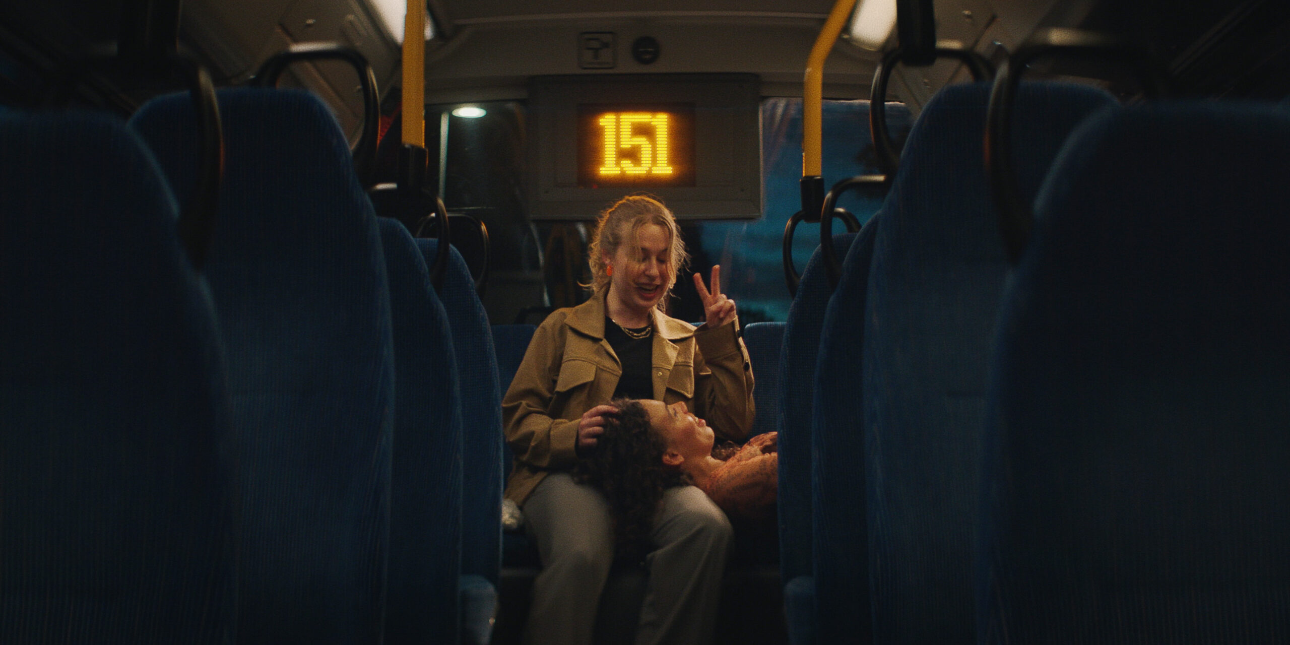 The interior of a bus at night looking down the aisle between rows of blue seats. In the back, a young woman in a tan jacket flashes a peace sign while another woman rests her head in her lap. An electronic sign overhead displays the number 151.