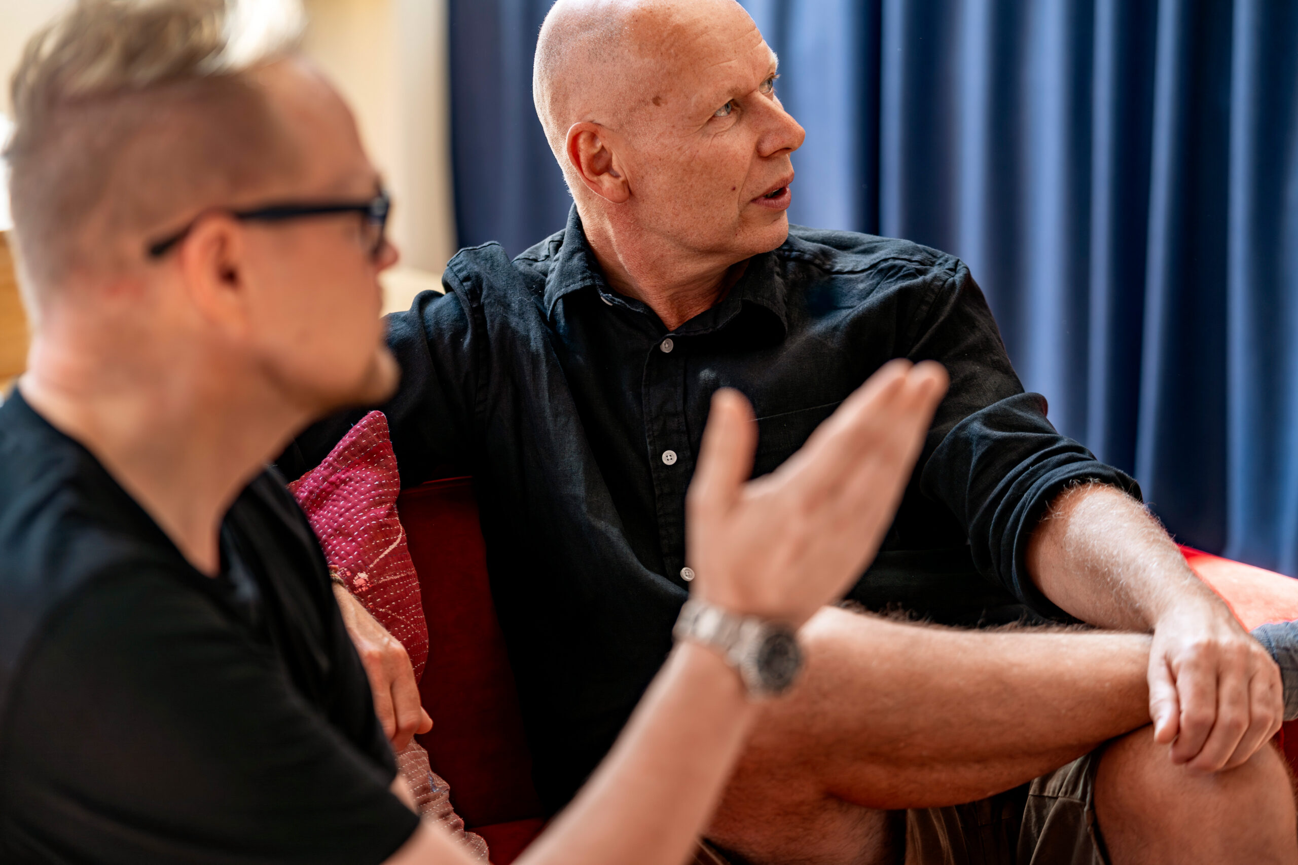 A bald man in a black button-down shirt sitting on a couch and listening intently as another person gesticulates with their hand in the foreground.