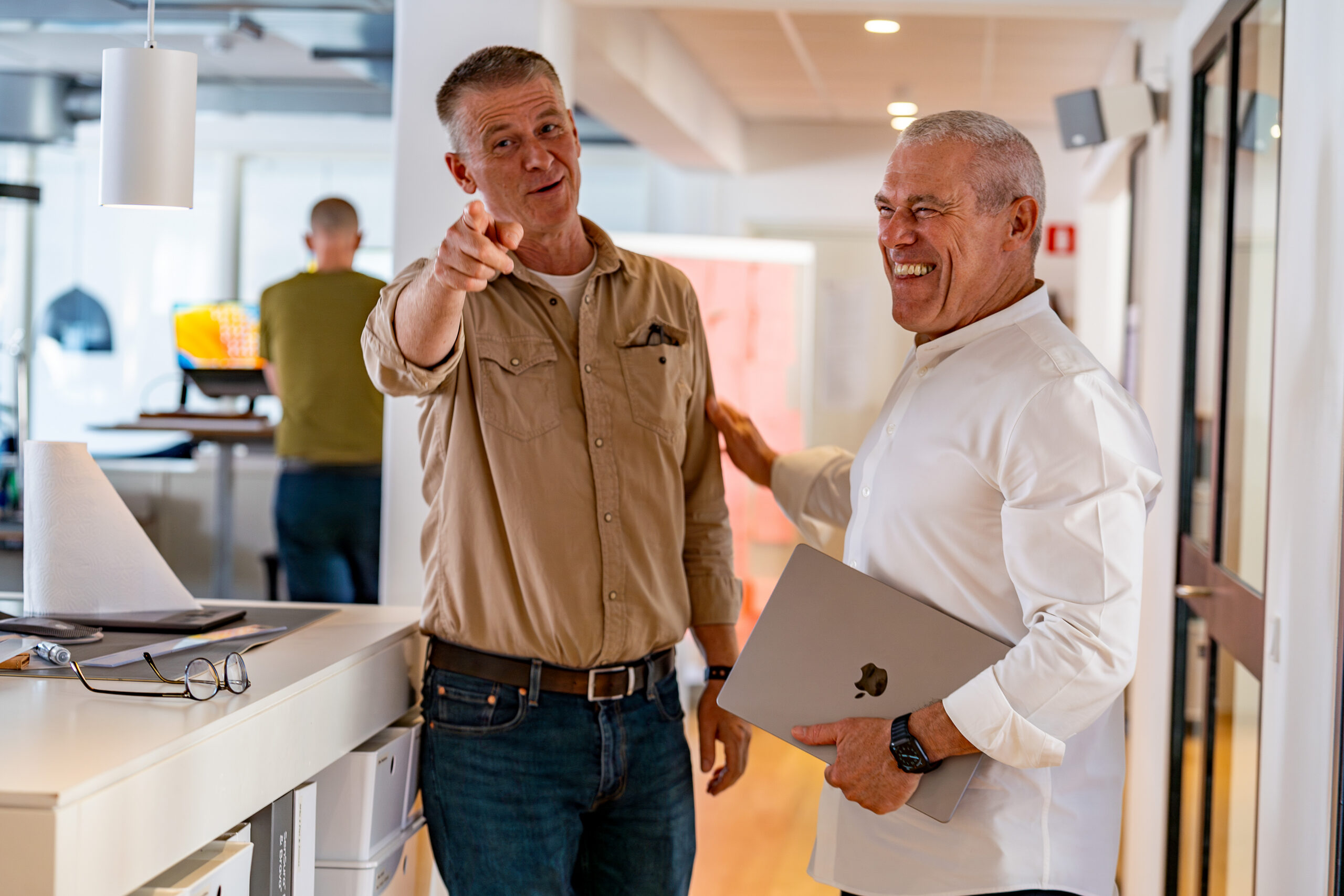 A man in a tan button-down shirt and jeans stands in a modern office hallway, smiling and pointing directly at the camera, while a laughing colleague in a white shirt stands next to him holding a closed laptop.