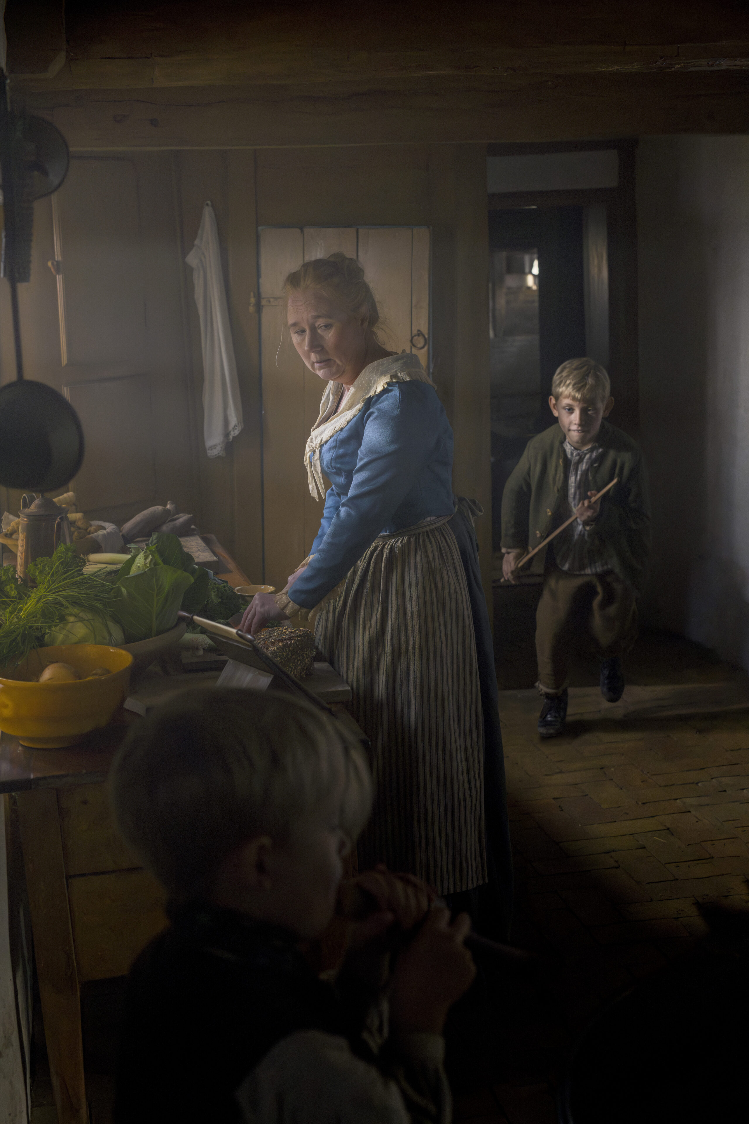 An indoor scene in a dimly lit, rustic historical kitchen. A woman in a blue 19th-century dress and striped apron prepares food at a counter, while a young boy runs into the room behind her with a wooden sword and another boy is seen in the foreground.