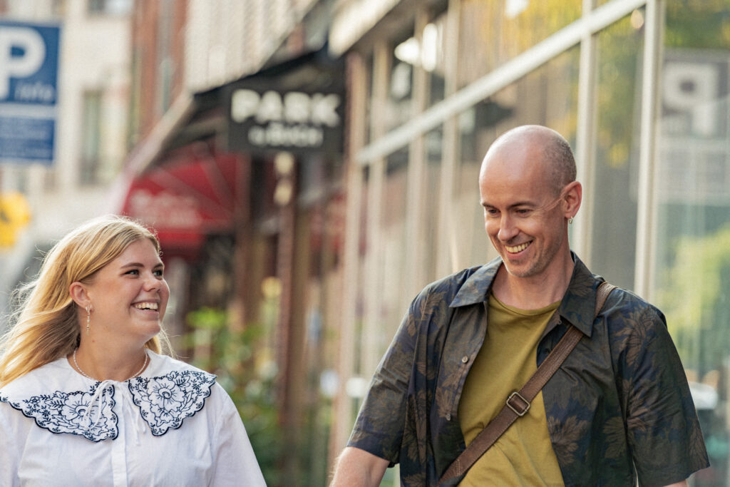 A blonde woman and a bald man walking down a city street together, smiling and laughing in a candid moment.