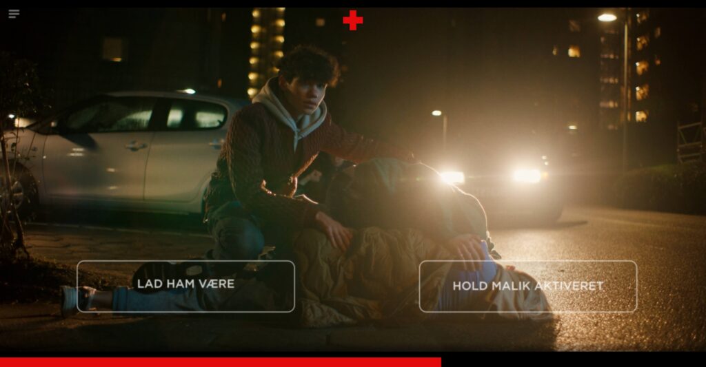 A scene from the interactive video where a young man kneels on a dark street beside a person lying down, with car headlights brightly shining behind them. Two interactive choice boxes are overlaid at the bottom: 'LAD HAM VÆRE' (Leave him alone) on the left, and 'HOLD MALIK AKTIVERET' (Keep Malik active) on the right.