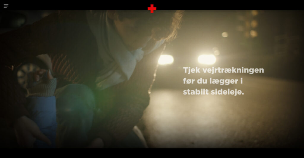 A video still from the first aid simulation showing a person kneeling over a casualty in the glare of car headlights at night. White instructional text overlay on the right reads: 'Tjek vejrtrækningen før du lægger i stabilt sideleje.' (Check the breathing before placing in the recovery position).
