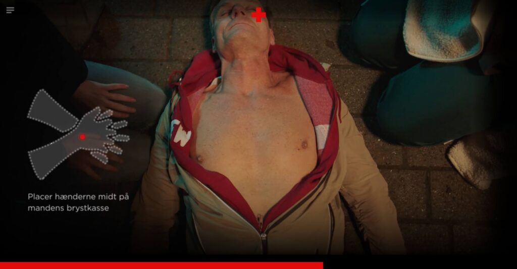 An overhead view within the interactive video showing an unconscious man lying on his back with his shirt unzipped, exposing his chest. On the left side, an animated icon of two hands appears over a red dot, accompanied by the text instruction: 'Placer hænderne midt på mandens brystkasse' (Place your hands in the middle of the man's chest).
