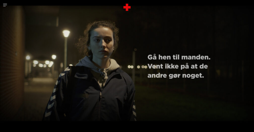 A video still showing a young woman in a navy track jacket walking down a dimly lit street at night. A large white text overlay on the right reads: 'Gå hen til manden. Vent ikke på at de andre gør noget.' (Go to the man. Don't wait for the others to do something).