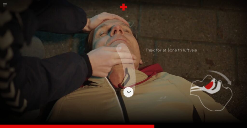 An interactive video frame showing a close-up of a person's hands tilting back the head of an unconscious man lying on the ground. A curved white interactive arrow icon is overlaid on his neck with the text: 'Træk for at åbne fri luftveje' (Pull to open the airway), alongside a anatomical diagram icon of a cleared throat.