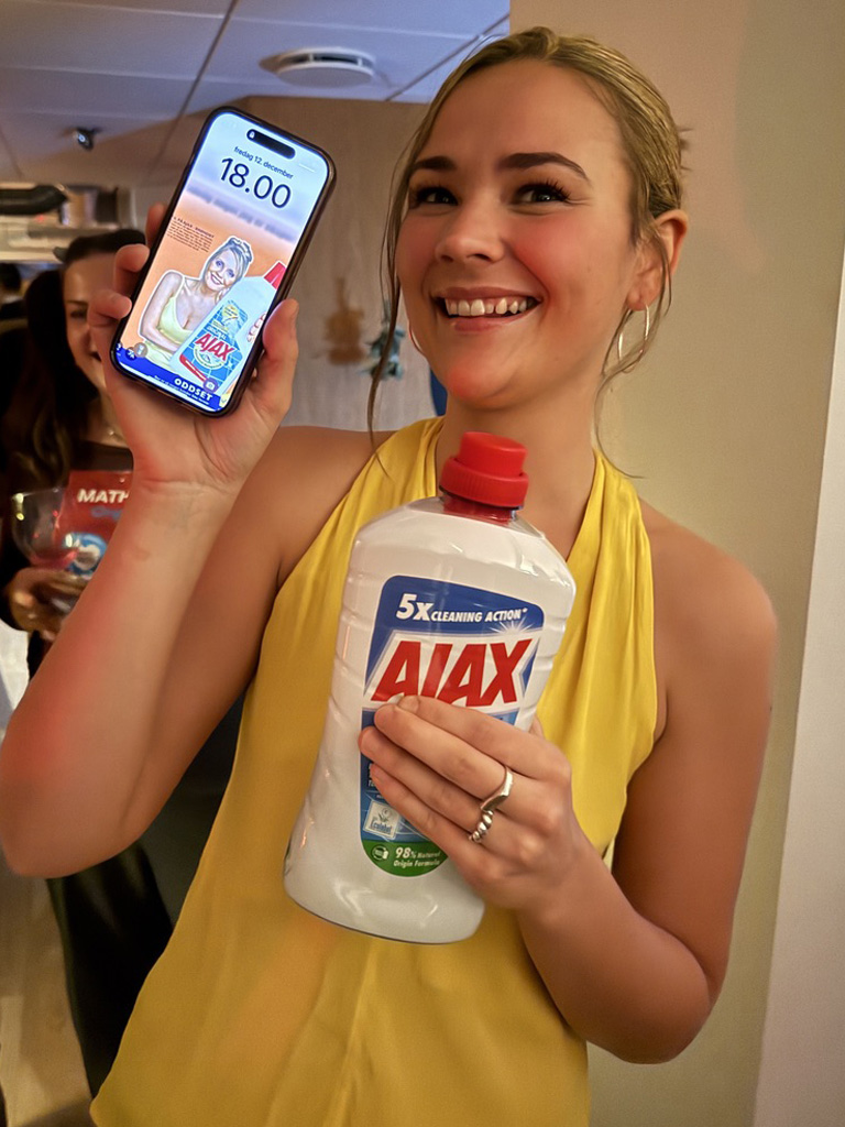 A smiling woman in a yellow top holding up a white bottle of Ajax cleaning solution in one hand and a smartphone showing a matching Ajax graphic in the other.