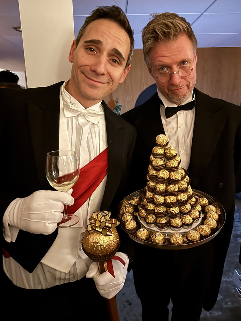 Two men dressed in formal black tuxedos and bow ties posing together; one holds a white wine glass and a large golden chocolate truffle, while the other carries a silver tray stacked with a pyramid of Ferrero Rocher chocolates.