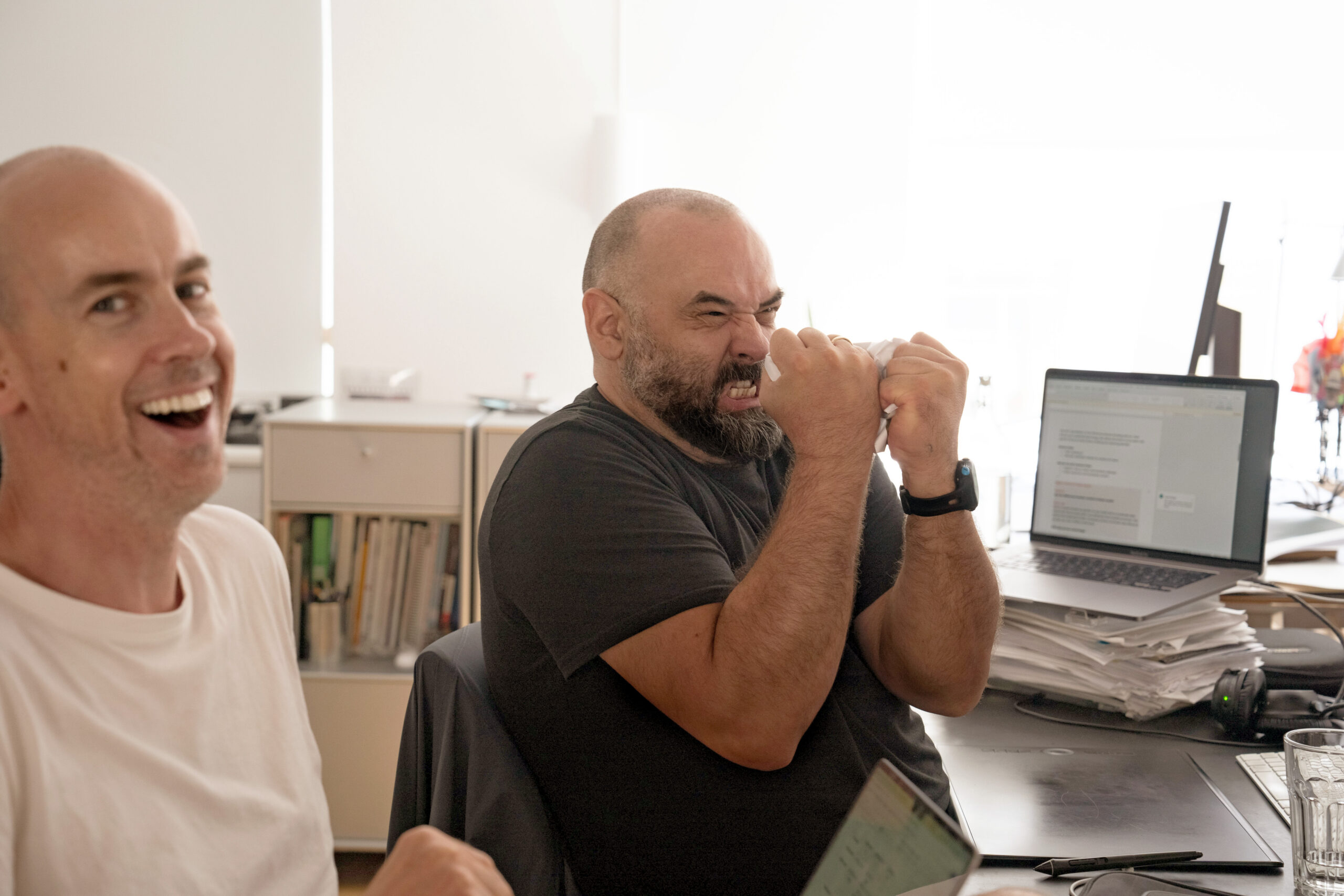 A man with a beard sits at an office desk, making a tense, frustrated face while clenching a piece of paper in his hands, while another man next to him laughs at the camera.