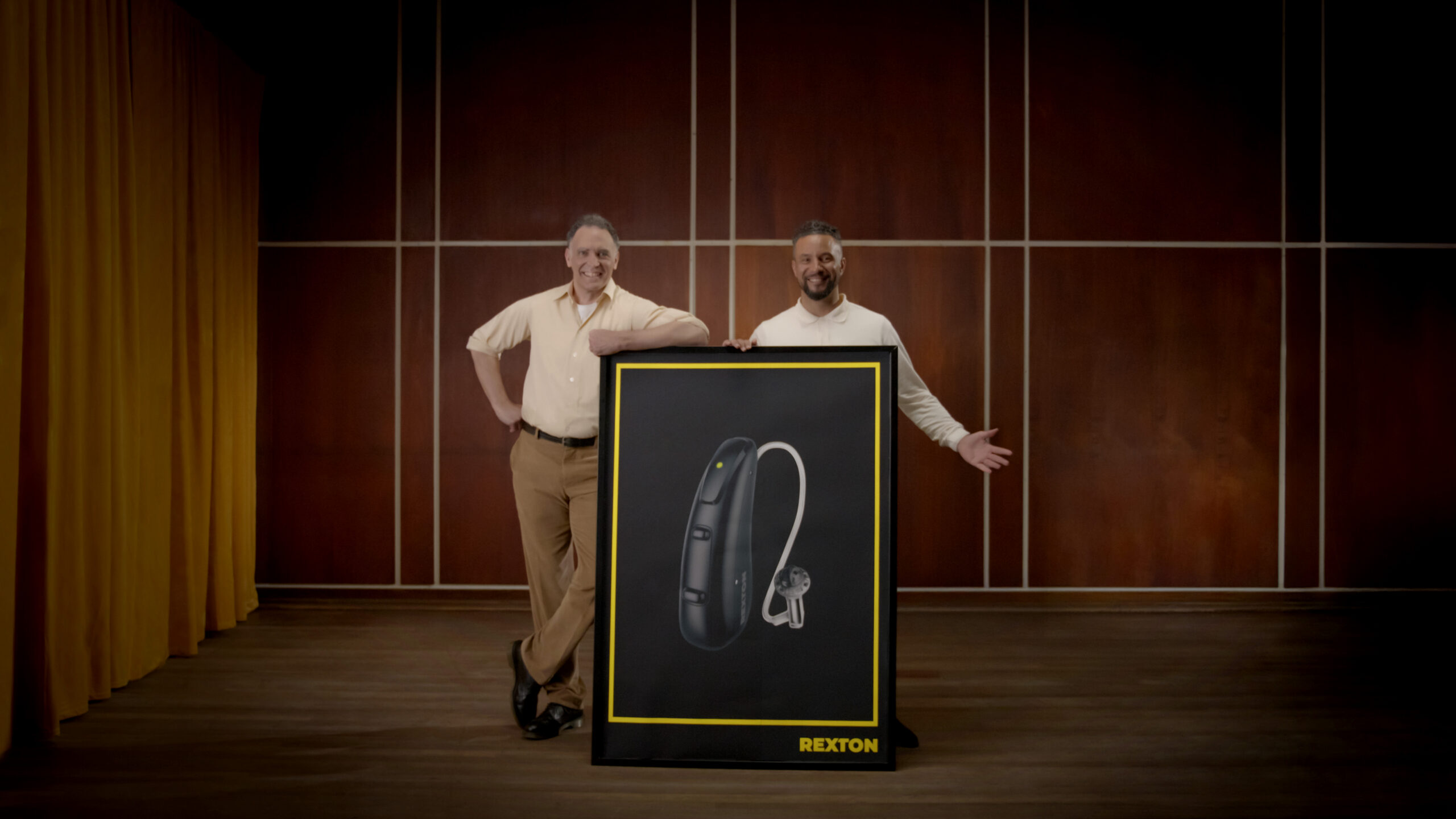 Two smiling men stand in a studio room with wood paneling and gold curtains, posing beside a large framed poster. The poster displays a close-up product shot of a black Rexton hearing aid against a black background.