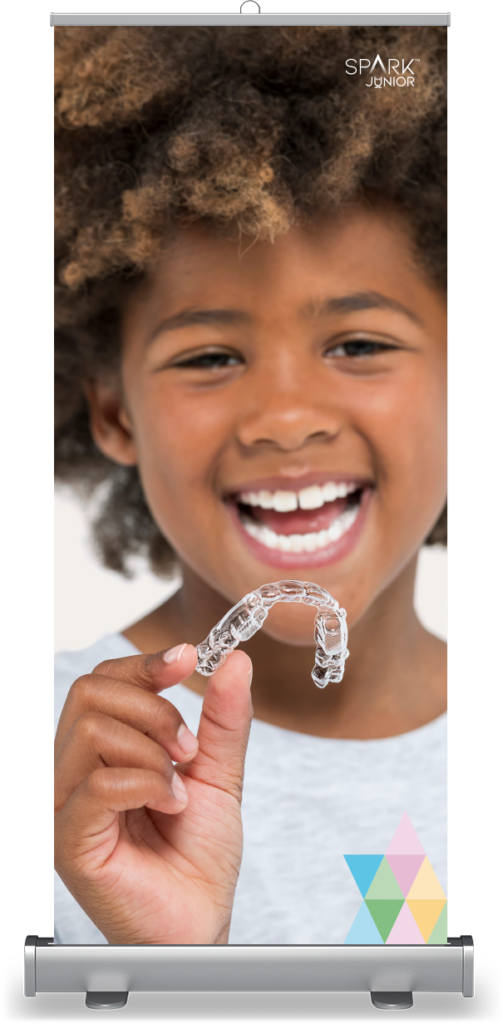 A rollup banner mockup featuring a close-up photo of a smiling young boy with curly hair. He is holding up a clear plastic dental aligner in front of his open, happy smile. The top right corner shows the 'SPARK JUNIOR' logo, and a colorful geometric pattern decorates the bottom right.