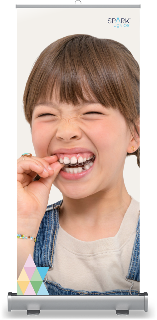 A rollup banner mockup featuring a young girl with braided pigtails winced playfully as she places a clear orthodontic retainer onto her teeth. The banner displays the 'SPARK JUNIOR' logo at the top right and a colorful triangular pattern in the bottom left corner.