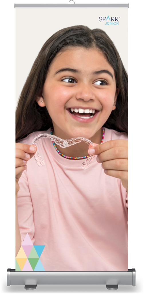 A rollup banner mockup featuring a young girl with long dark hair, smiling excitedly while holding a clear plastic dental aligner with both hands. The 'SPARK JUNIOR' logo is in the top right corner, and a colorful pastel triangular pattern sits at the bottom left.