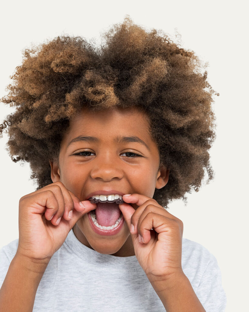 A close-up portrait of a smiling young boy with an afro hairstyle using his fingers to insert a clear plastic dental aligner onto his top teeth, looking directly at the camera against a light grey background.